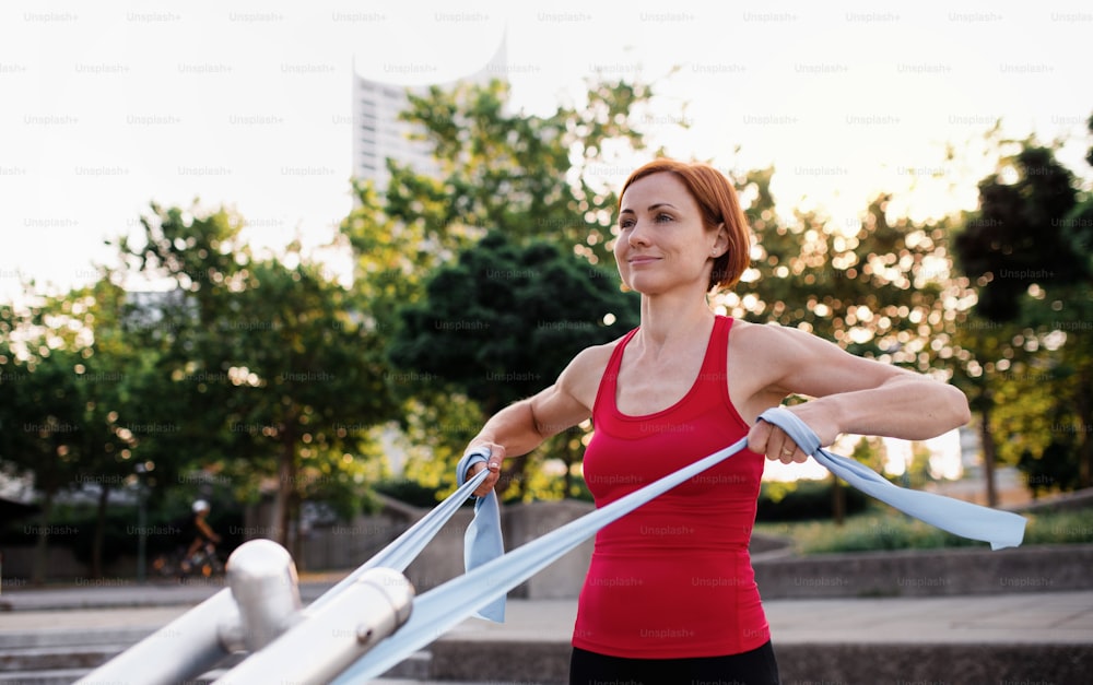 Full length portrait of young woman doing exercise outdoors in city with elastic bands.