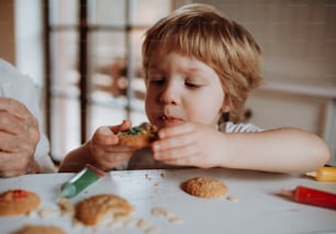Un niño pequeño y feliz sentado a la mesa, decorando y comiendo pasteles en casa.