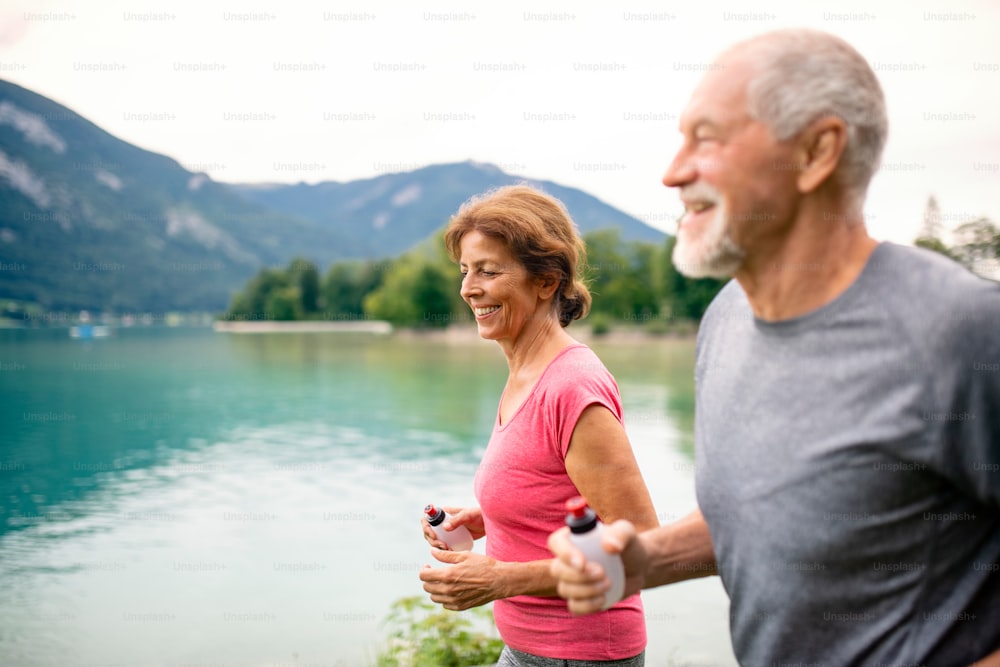 A senior pensioner couple with smartphone running by lake in nature.