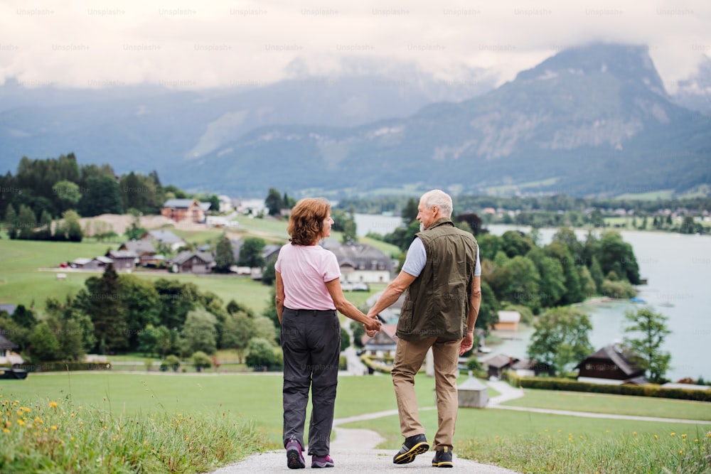 A rear view of senior pensioner couple hiking in nature, holding hands.