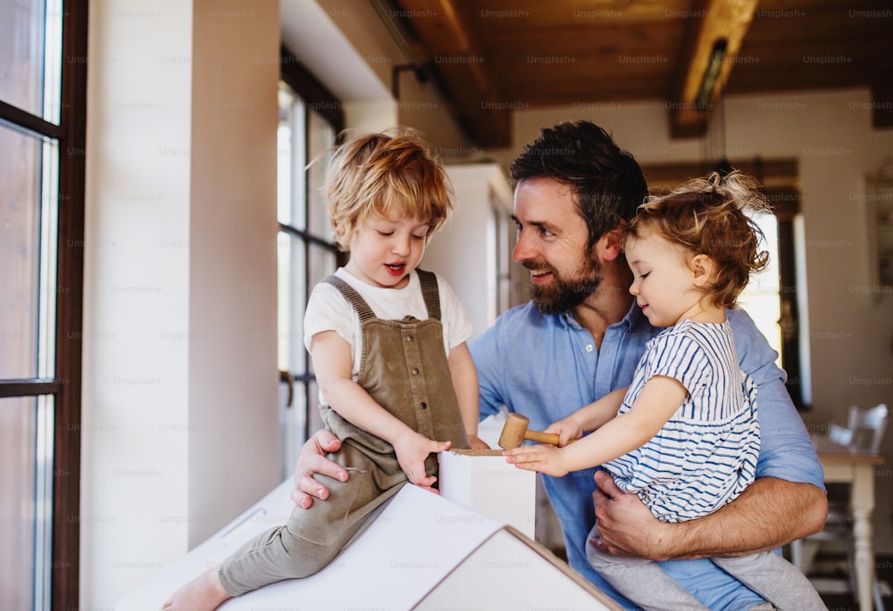 Deux enfants en bas âge heureux avec leur père jouant avec une maison en papier à l’intérieur à la maison.