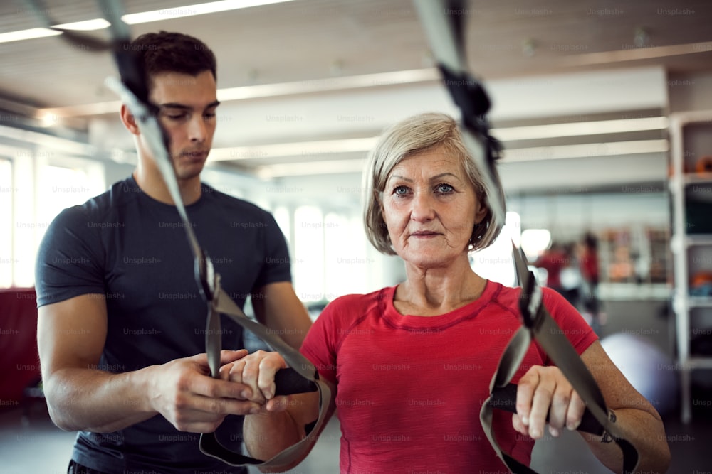 A senior woman in gym with a young personal trainer doing exercise with TRX.