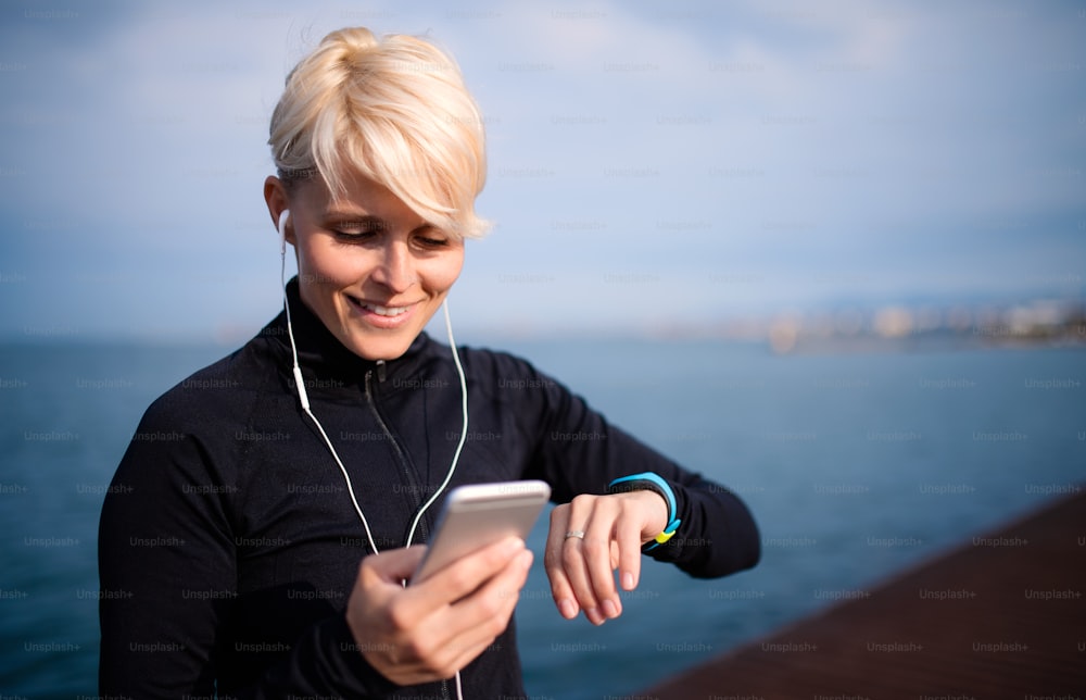 A midsection of young sportswoman with earphones standing outdoors on beach, using smartwatch and smartphone.