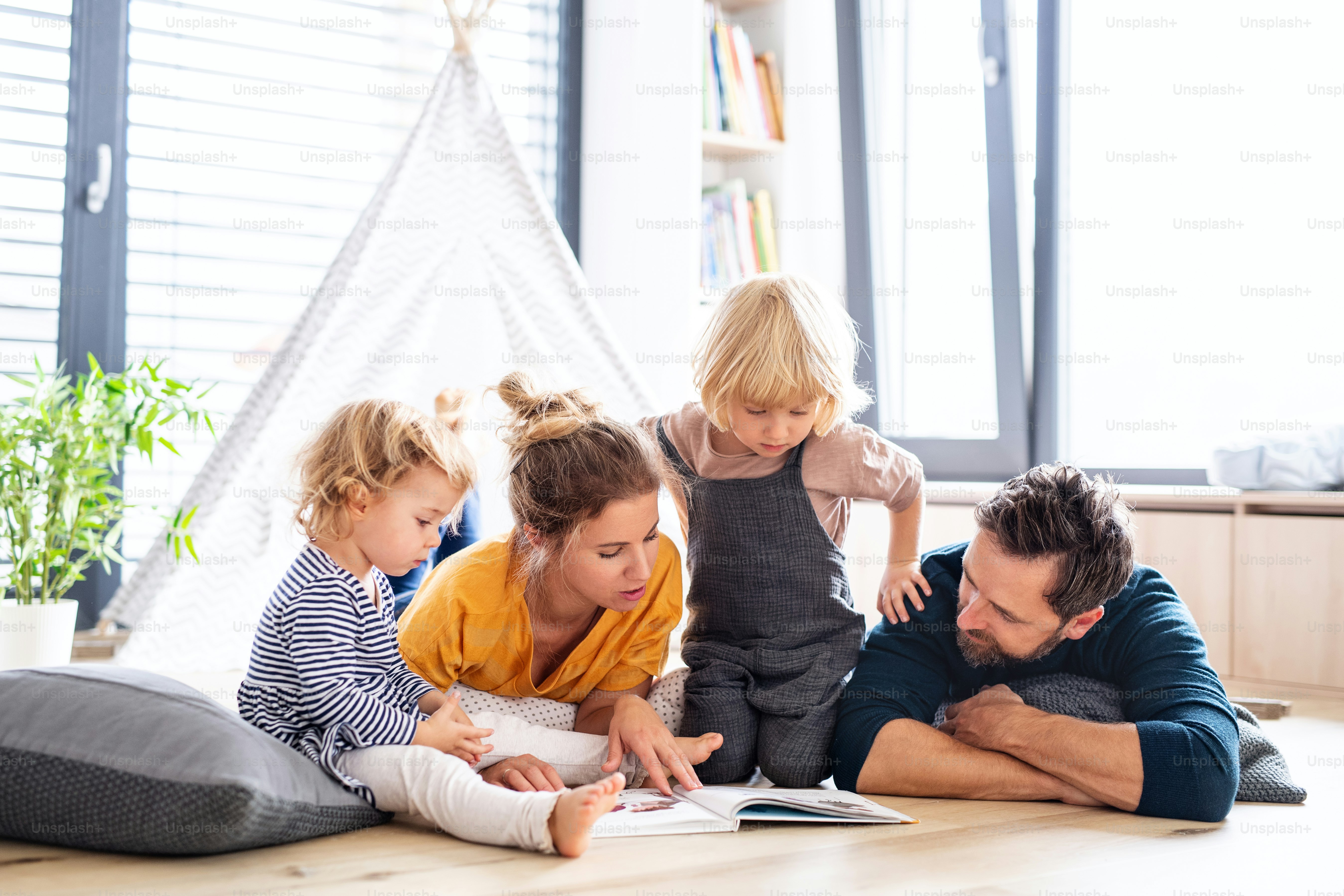 Vorderansicht der jungen Familie mit zwei kleinen Kindern drinnen im Schlafzimmer, die ein Buch lesen.