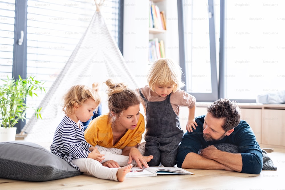 Vista frontal da família jovem com duas crianças pequenas dentro de casa no quarto lendo um livro.