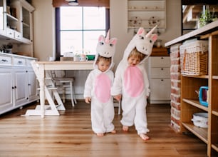 Dos niños pequeños con máscaras de unicornio blanco caminando por el interior de su casa.