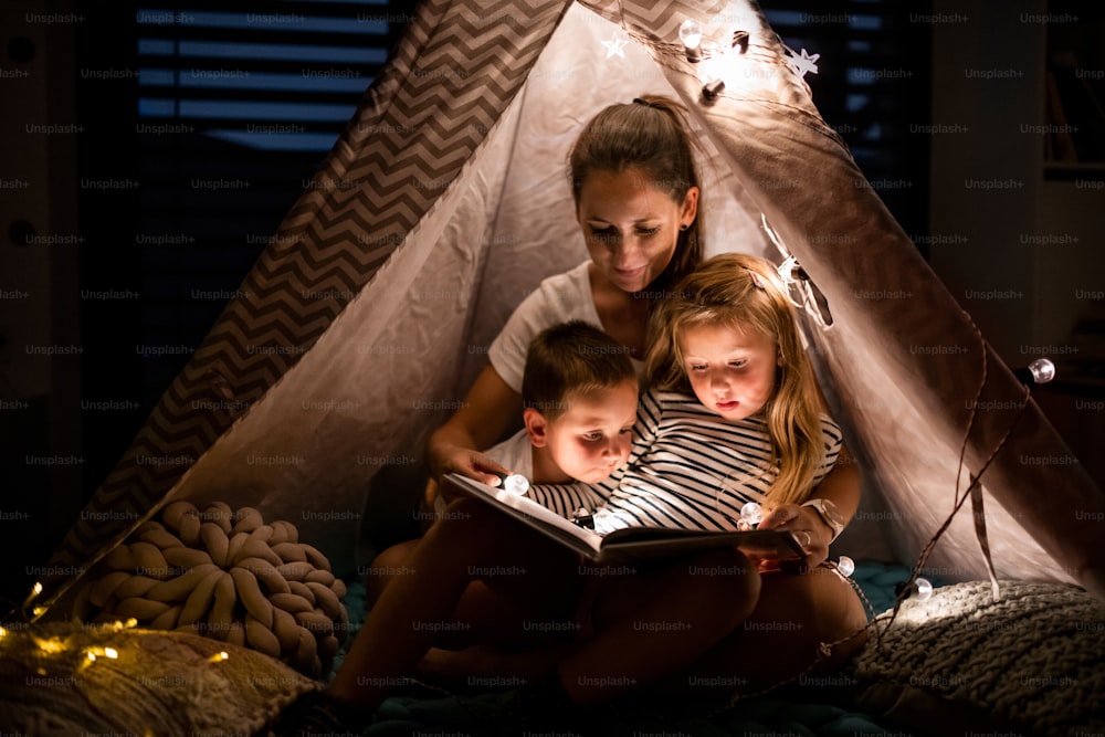 Mutter mit kleinen Kindern sitzt abends drinnen im Zelt im Schlafzimmer und liest ein Buch.