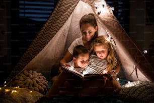 Madre con niños pequeños sentados en el interior en la tienda de campaña en el dormitorio por la noche, leyendo un libro.