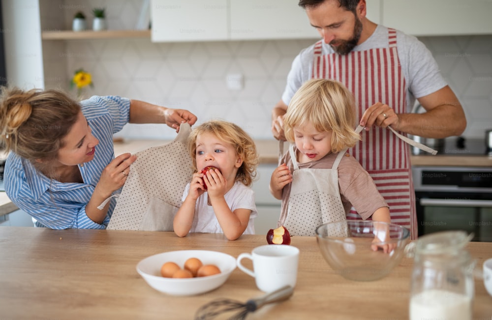 Vista frontal da família jovem com duas crianças pequenas dentro de casa na cozinha, cozinhando.