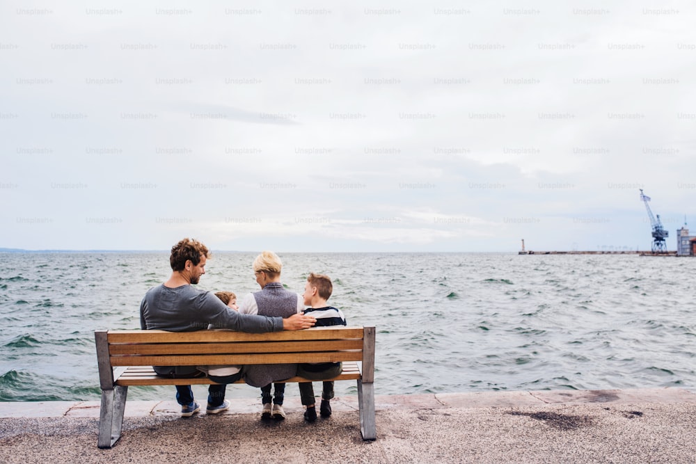 Rear view of young family with two small children sitting on bench outdoors on beach.