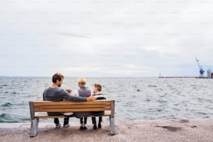 Rear view of young family with two small children sitting on bench outdoors on beach.