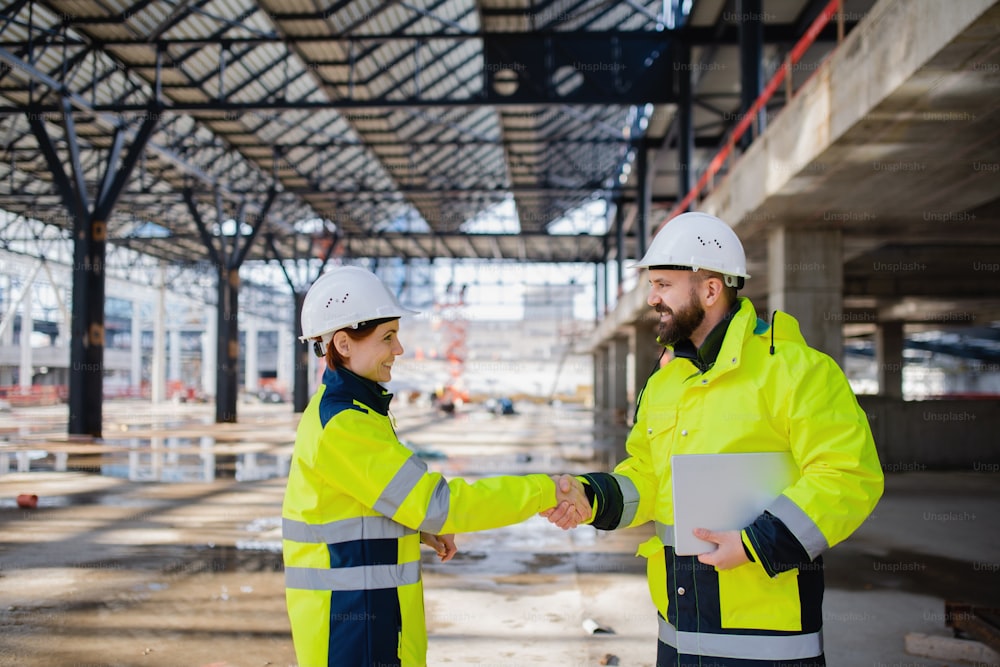 Two engineers with tablet standing on construction site, shaking hands.