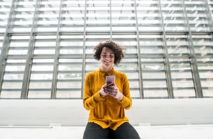 A portrait of a young student or businesswoman sitting on desk in room in a library or office, using smartphone.