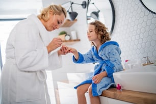 A happy cute small girl with mother in bathroom indoors at home, painting nails.