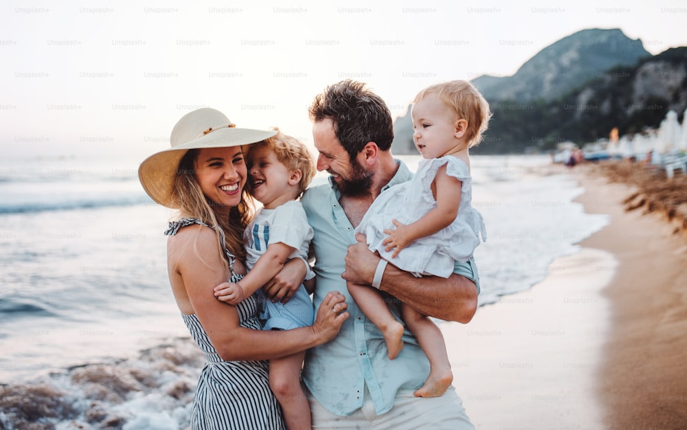 Una giovane famiglia con due bambini piccoli in piedi sulla spiaggia durante le vacanze estive, ridendo.