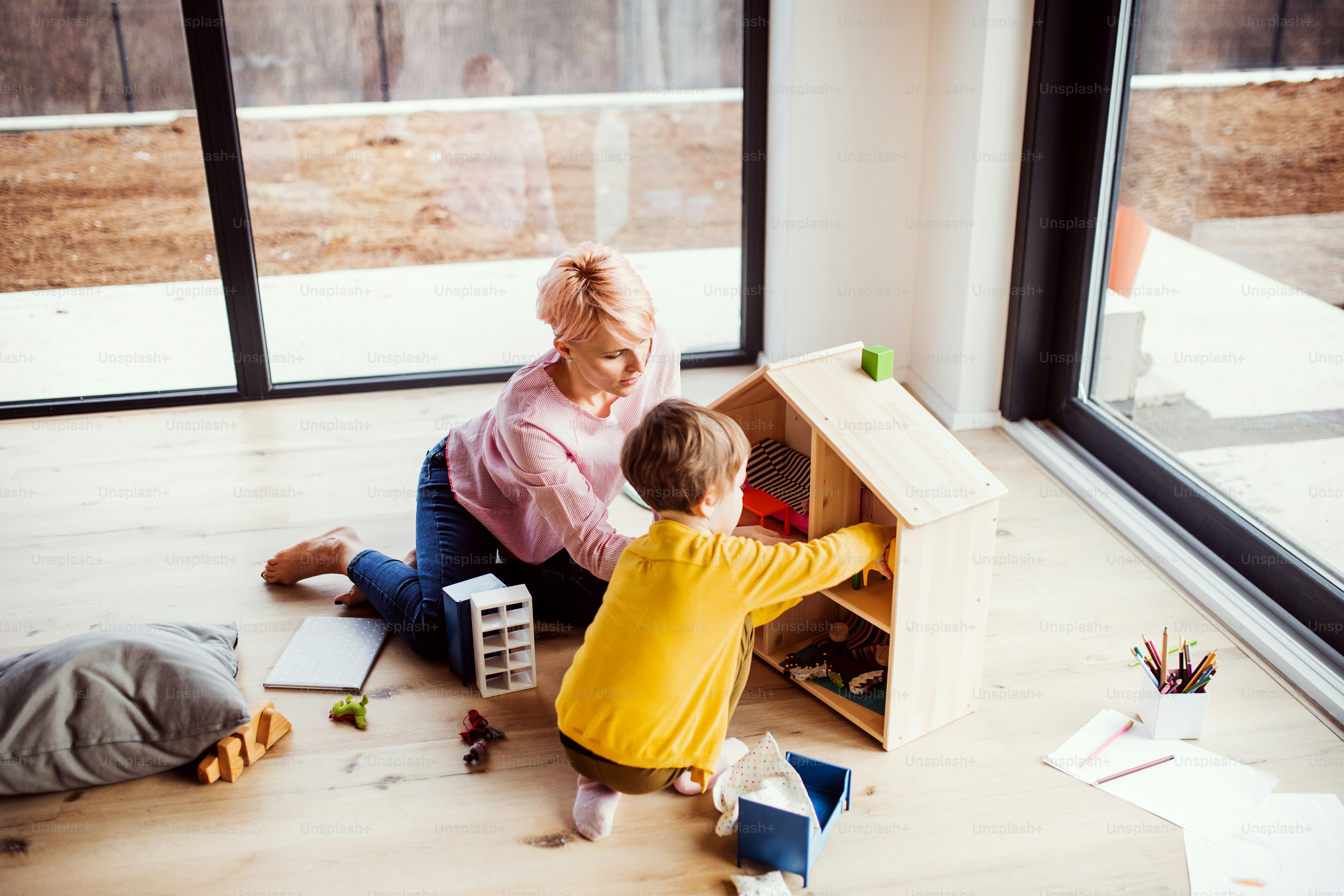 A young woman and small daughter playing with wooden house on the floor, top view.