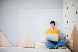 Front view of young woman with laptop sitting on bed in bedroom indoors at home. Copy space.