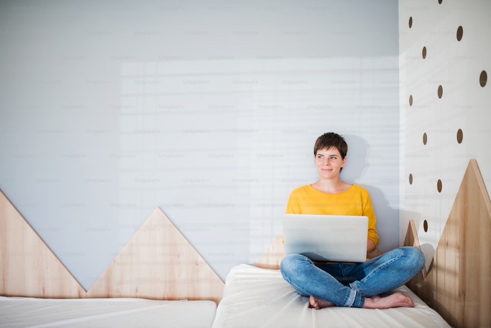 Vista frontal da mulher nova com laptop sentado na cama no quarto dentro de casa. Espaço de cópia.