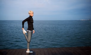 Side view portrait of young sportswoman standing outdoors on beach, stretching. Copy space.