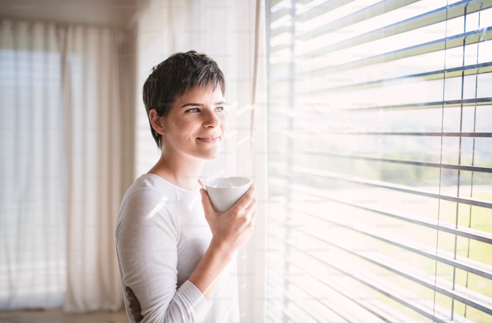 Portrait of young happy woman with coffee standing by window indoors at home.