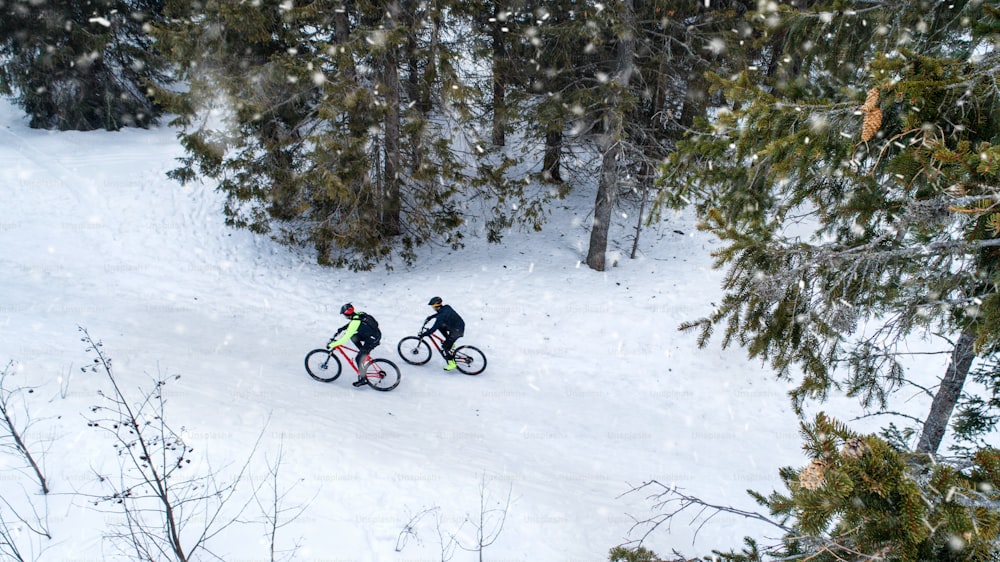 Aerial view of two mountain bikers riding on road covered by snow in forest outdoors in winter.