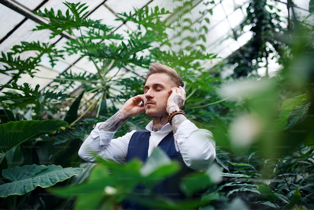 Junger Mann mit Kopfhörern steht im botanischen Garten und hört Musik. Green Businessness-Konzept.