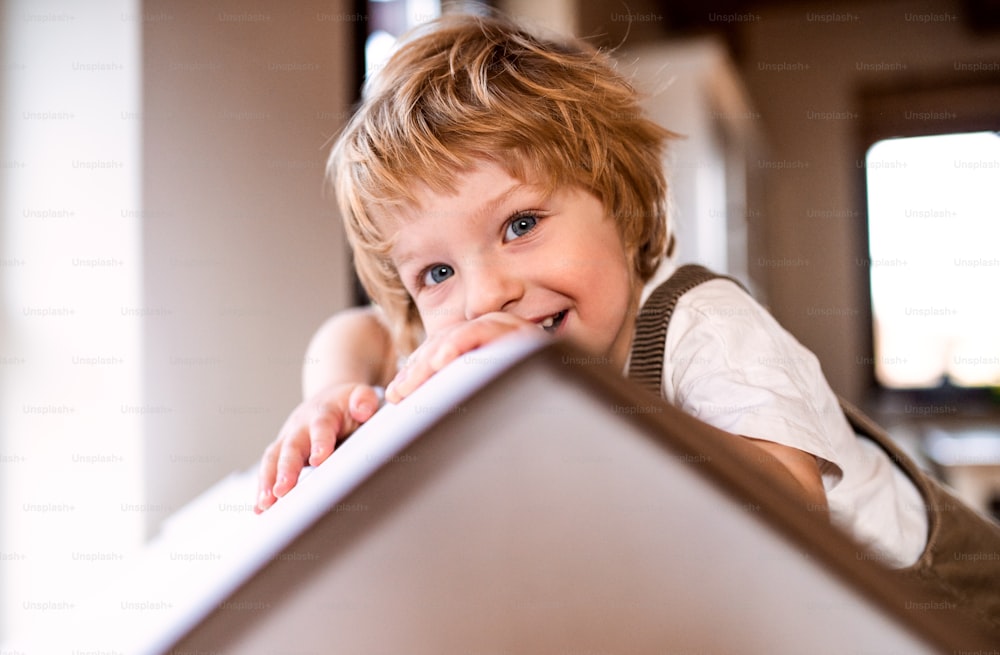 A happy toddler boy playing with a carton paper house indoors at home.