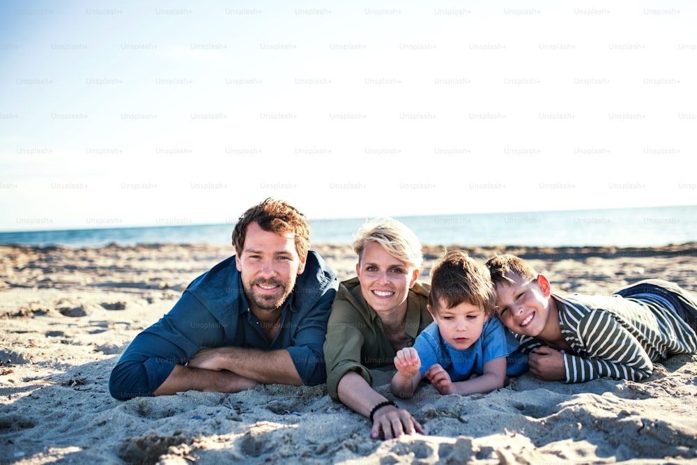 Retrato de una familia joven con dos niños pequeños acostados al aire libre en la playa, mirando a la cámara.