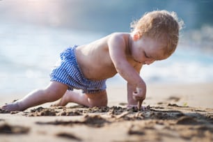 A small topless toddler girl on beach on summer holiday, playing in sand.