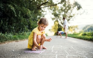 晴れた夏の自然の中、田舎道でチョークで絵を描いている小さなかわいい女の子。