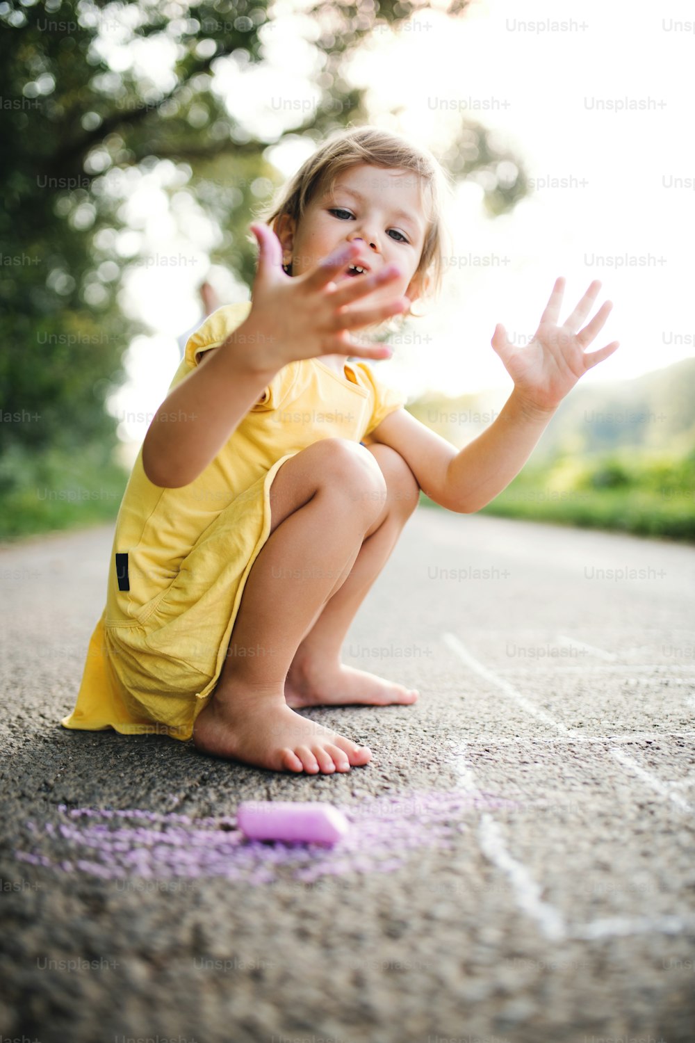 A small cute girl on a road in countryside in sunny summer nature, drawing with chalk.