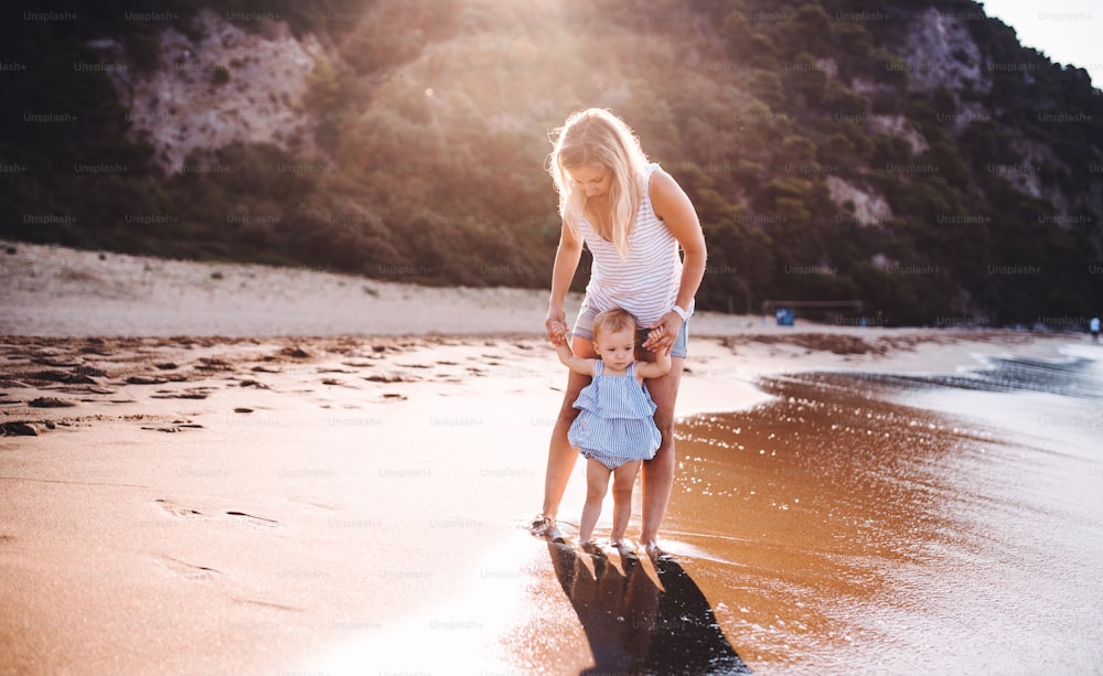 Madre joven caminando con una niña pequeña en la playa en vacaciones de verano al atardecer.
