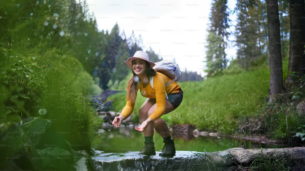 Happy young woman standing by stream on a walk outdoors in summer nature, looking at camera.