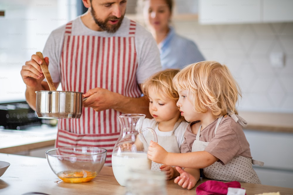 Uma jovem família com duas crianças pequenas dentro de casa na cozinha, cozinhando.