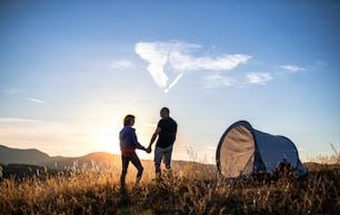 A rear view of senior tourist couple with shelter tent standing in nature at sunset, resting.