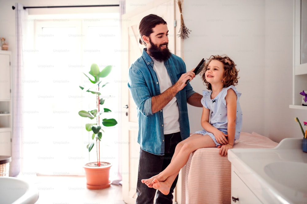 Un padre joven cepillando el cabello de una hija pequeña feliz en el baño de su casa.
