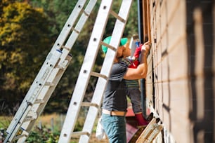 A side view of father and toddler boy outdoors in summer, painting wooden house.