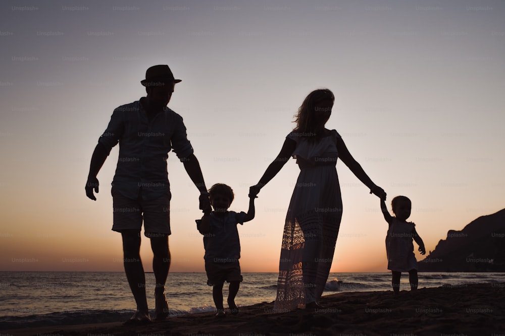 A family with two toddler children walking on beach on summer holiday at dusk, silhouettes holding hands.