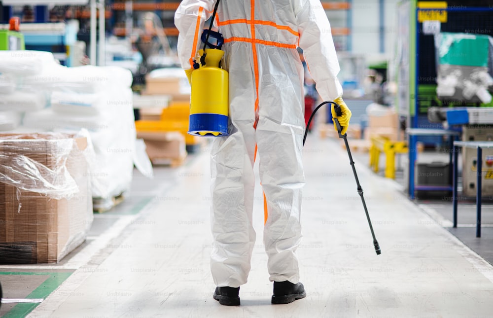 Rear view portrait of man worker with protective mask and suit disinfecting industrial factory with spray gun.
