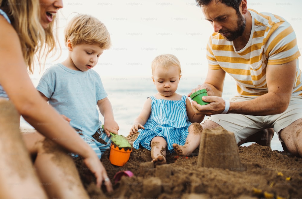 A young family with toddler children playing with sand on beach on summer holiday.