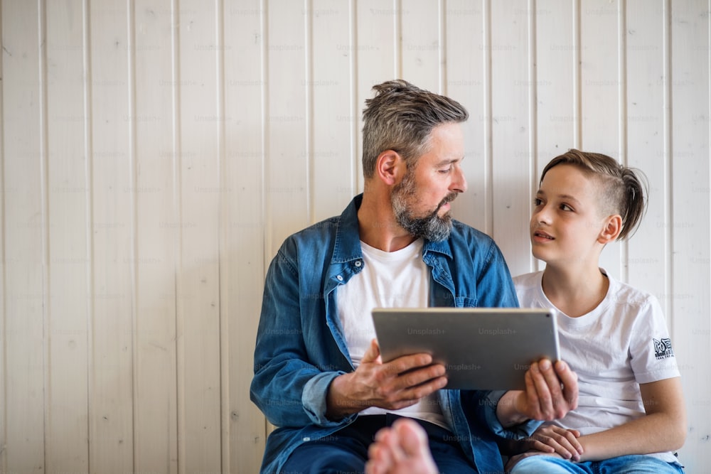 Mature father with small son sitting indoors against white wall, using tablet. Copy space.