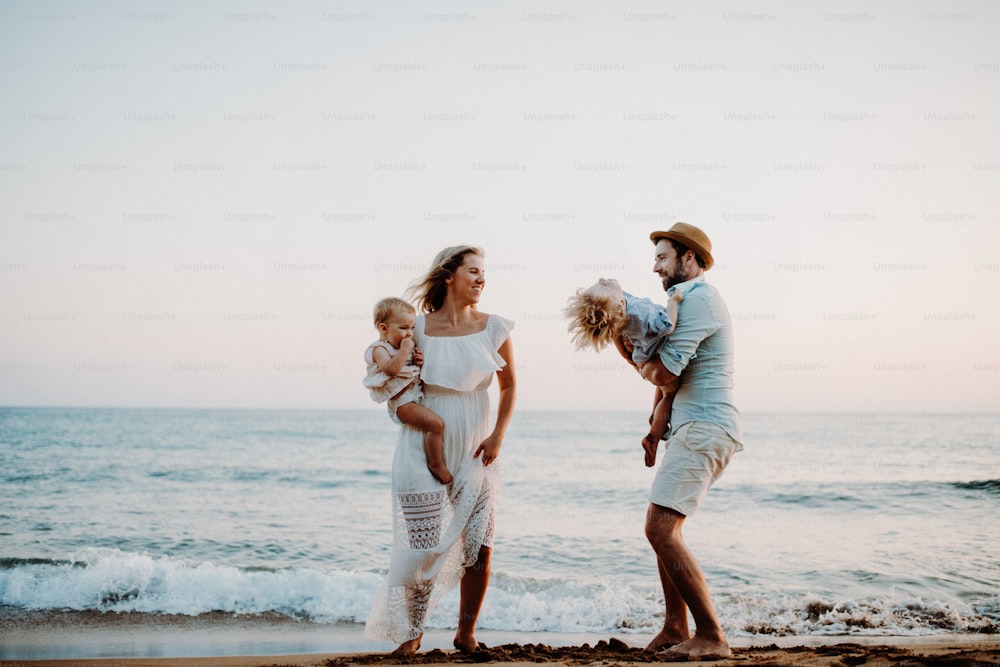 Una familia joven con dos niños pequeños de pie en la playa durante las vacaciones de verano, riendo.