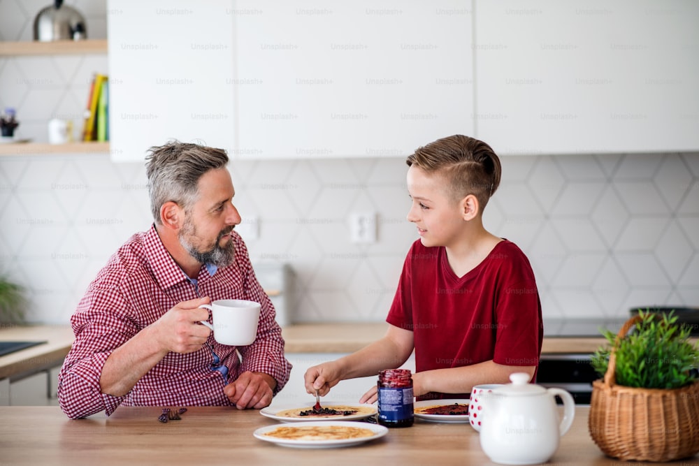 A mature father with small son indoors sitting at the table, making and eating pancakes.