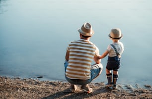 A rear view of father with a small toddler son spending time outdoors by a lake. Copy space.
