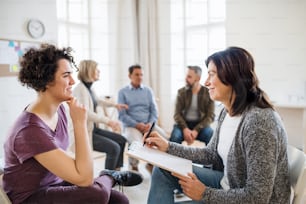 A senior counsellor with clipboard talking to a young woman during group therapy.