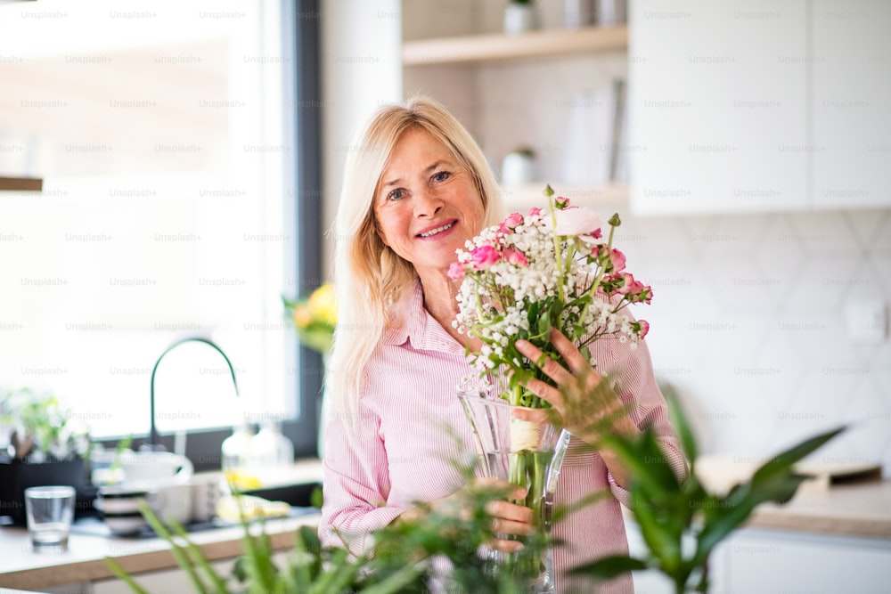 Portrait of happy senior woman arranging flowers in vase indoors at home.