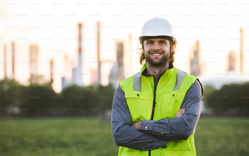 Retrato de un joven ingeniero de pie al aire libre junto a la refinería de petróleo, mirando a la cámara.