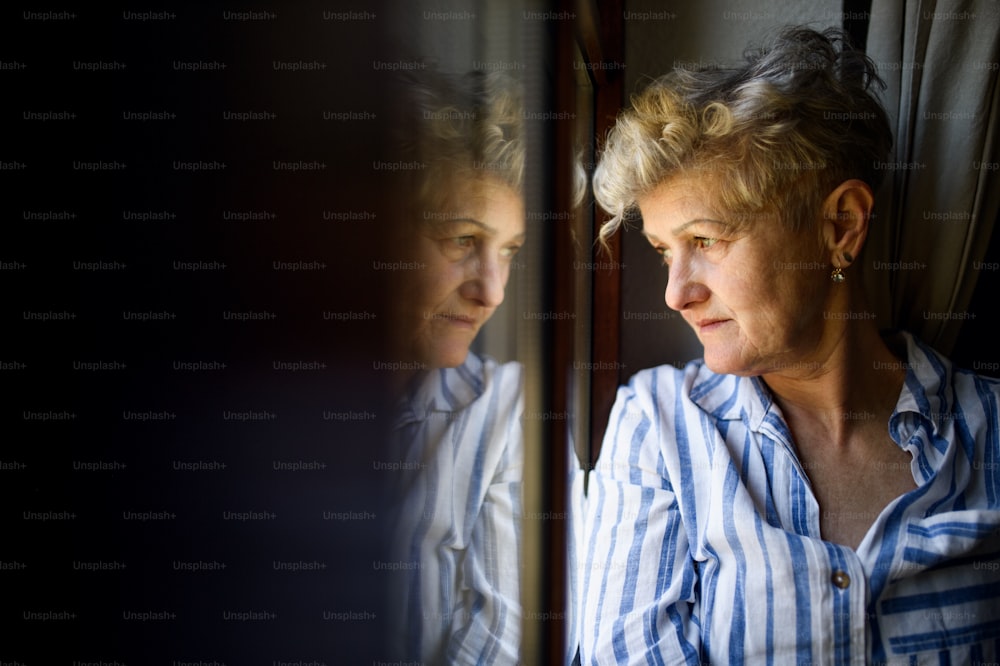 自宅の窓際に屋内に立っている悲しい年配の女性、コロナウイルスと検疫のコンセプト。スペースをコピーします。