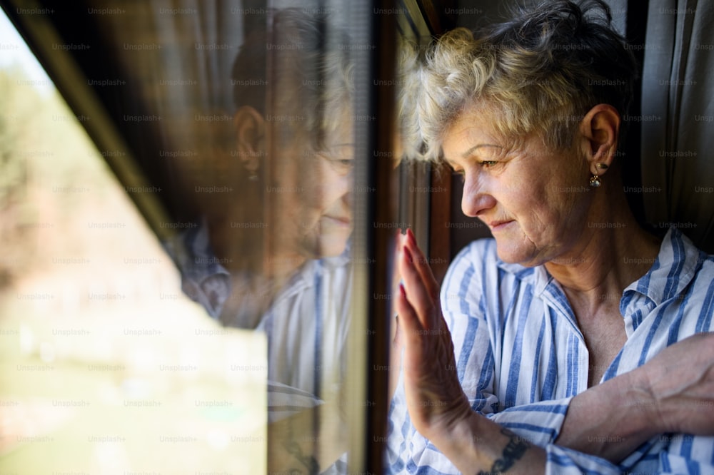집에서 창문 옆에 실내에 서 있는 슬픈 노인 여성, 코로나 바이러스 및 격리 개념.