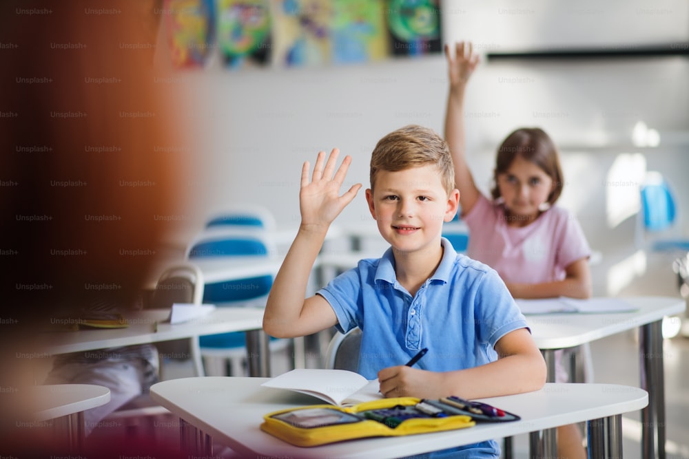 Niños pequeños de la escuela sentados en el escritorio en el aula en la lección, levantando las manos.